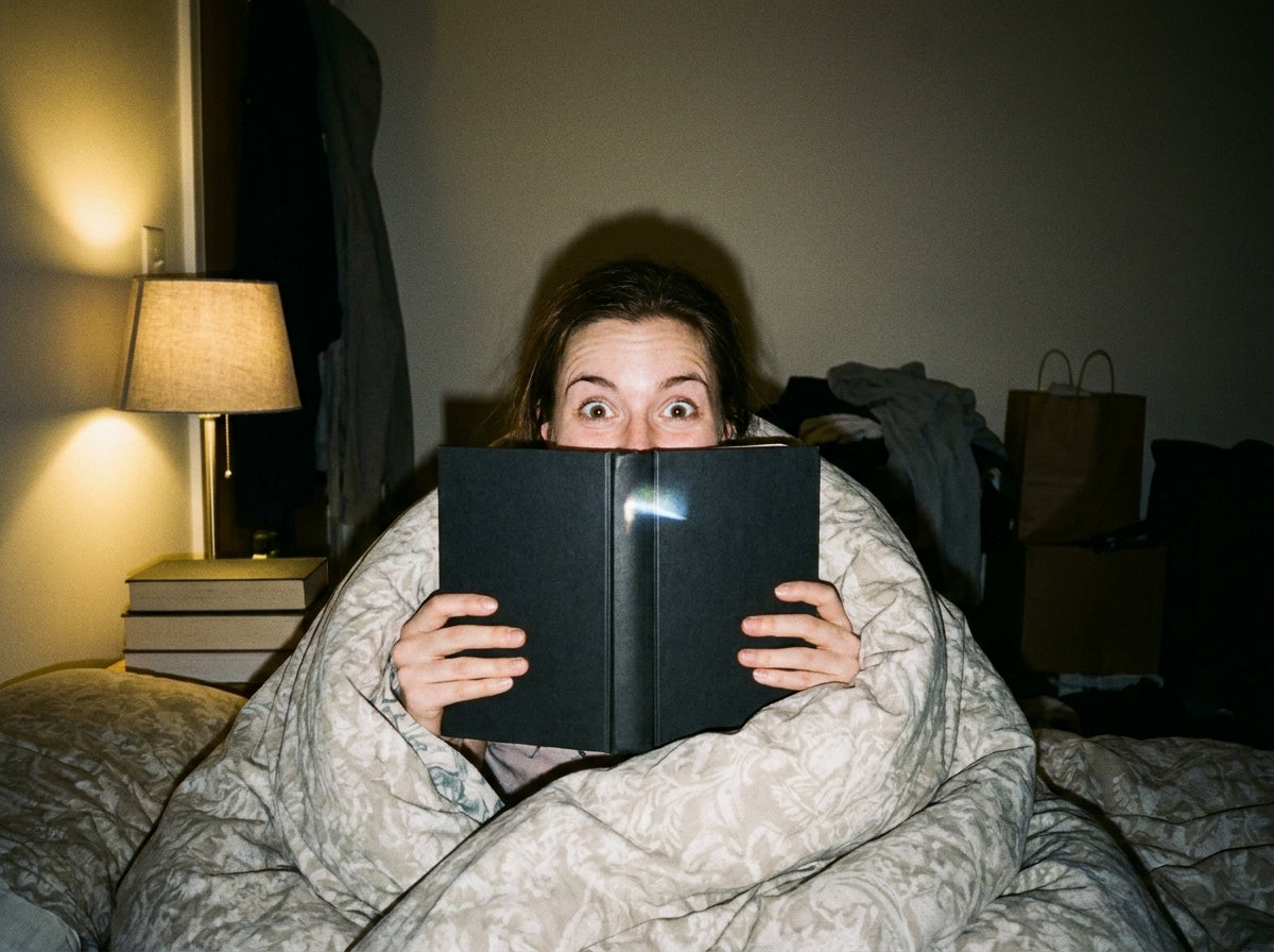 A reader wrapped in a duvet, wide-eyed over the top of a dark hardcover book in bed at night