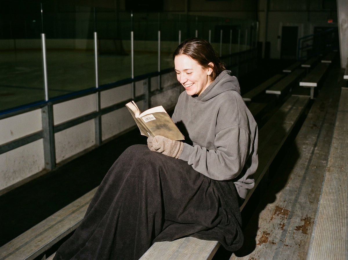 A reader wrapped in a blanket on the bleachers of a cold ice rink, smiling at her book